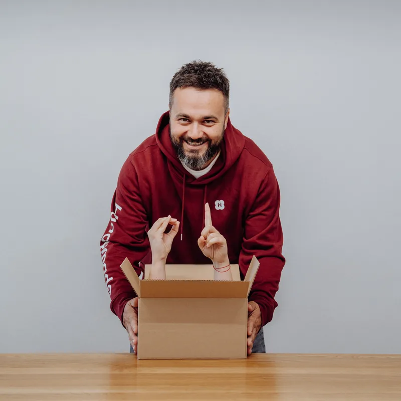Aus einem geöffneten Karton ragen zwei Hände, dahinter lacht ein Teammitglied im bordeauxroten Handymade-Hoodie