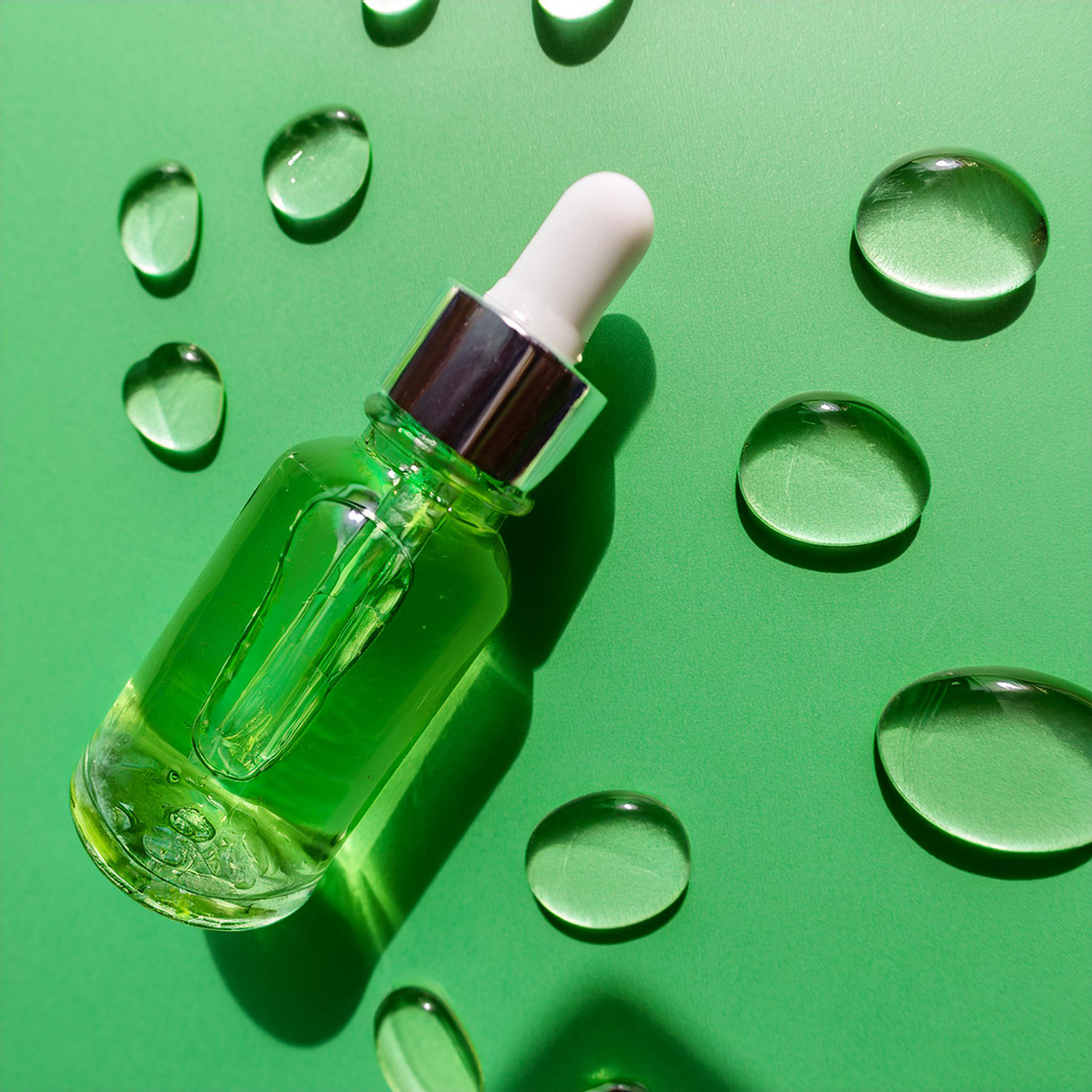 Blick auf ein Glasfläschchen mit Pumpverschluss, gefüllt mit einer transparenten Flüssigkeit – Hyaluronsäure-Serum. Um das Fläschchen mit dem Serum herum befinden sich mehrere Tropfen des verschütteten Serums auf dem Hintergrund. Den Hintergrund bildet ein einfarbiger, zart grüner Hintergrund. Das Fläschchen mit dem Serum liegt. Blick von oben und Studioausleuchtung